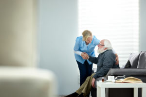 Female caregiver helping senior elderly man get up from couch at assisted living memory care